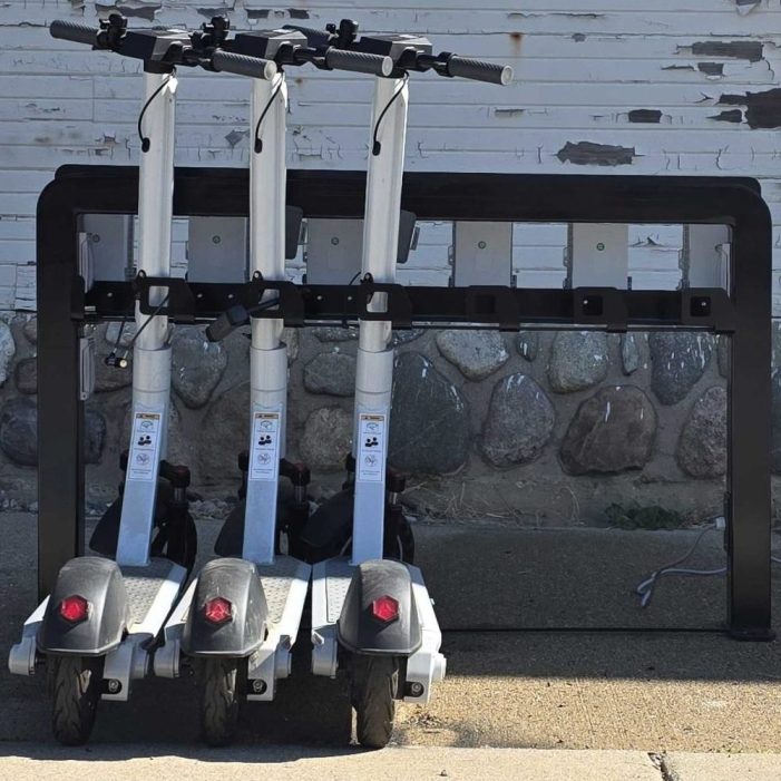 Three e-scooters docked in a Juise charging rack against a stone wall