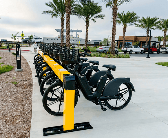 Row of e-bikes docked at a yellow Juise charging station in a palm-lined plaza