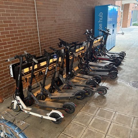 Electric scooters parked in a Juise charging rack against a brick wall outside a college residence hall